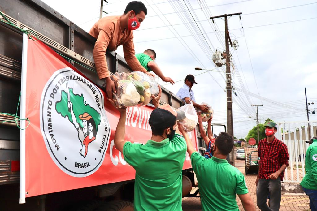 fotografia colorida mostra um caminhão estacionado e dele as pessoas descarregam cestas de alimentos para doação. no caminhão, há uma faixa com o símbolo do MST