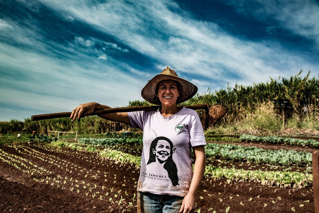 foto colorida em que no primeiro plano está uma mulher camponesa sorrindo e segurando no ombro uma enxada. ao fundo, aparece a horta cultivada e plantações